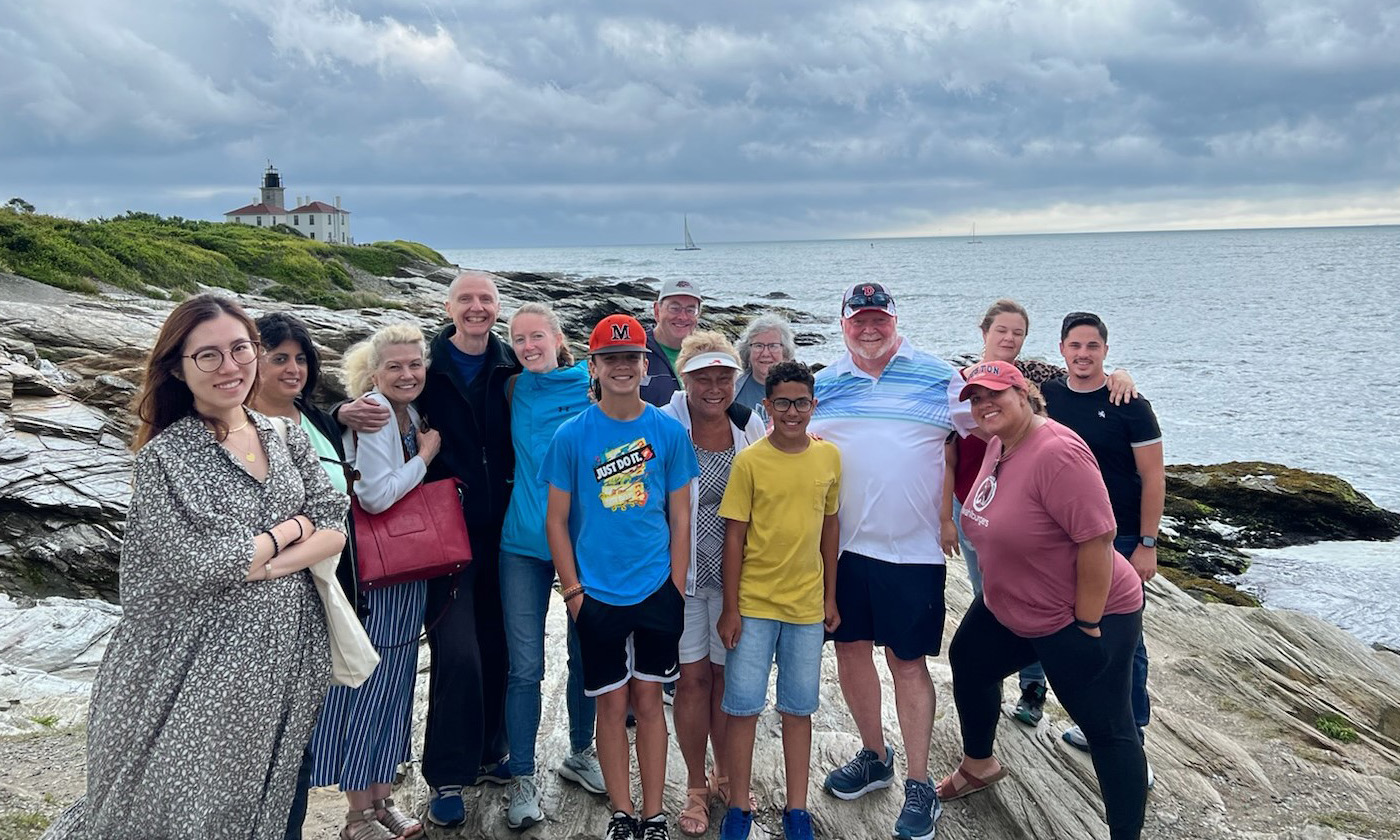 Group of people near the shore posing to the camera with the view of Beavertail Lighthouse, Jamestown, RI.